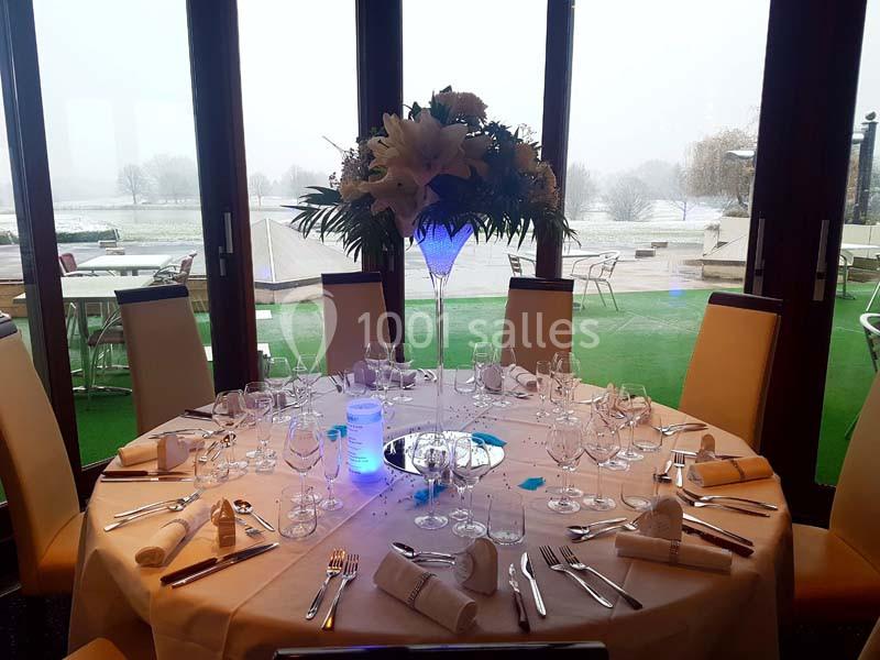 Table ronde élégamment dressée avec nappes blanches, verres et couverts, décorée d'un grand centre floral, vue sur jardin.