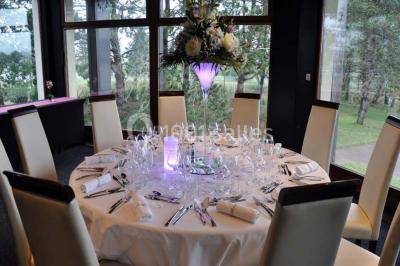 Salle de réception décorée avec des tables rondes, nappes blanches et chaises ornées de nœuds colorés.