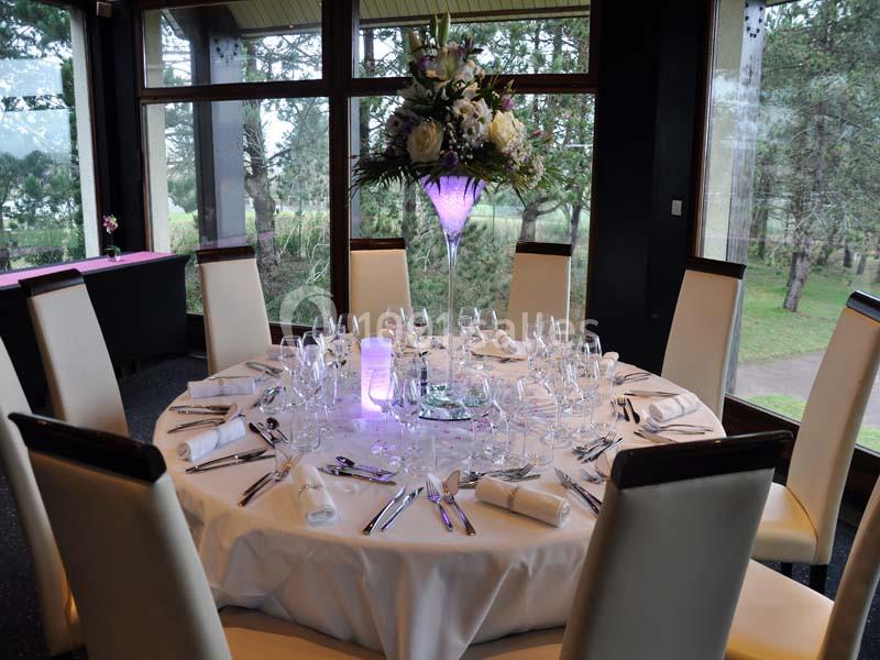 Table ronde élégamment dressée avec nappes blanches, verres, couverts et centre de table floral dans une salle lumineuse.