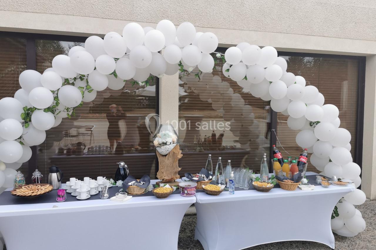 Buffet extérieur décoré avec une arche de ballons blancs, comprenant boissons, viennoiseries et collations variées.