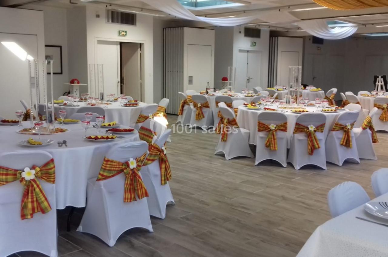 Salle décorée pour un événement, avec des tables rondes dressées et des chaises ornées de nœuds en tissu coloré.