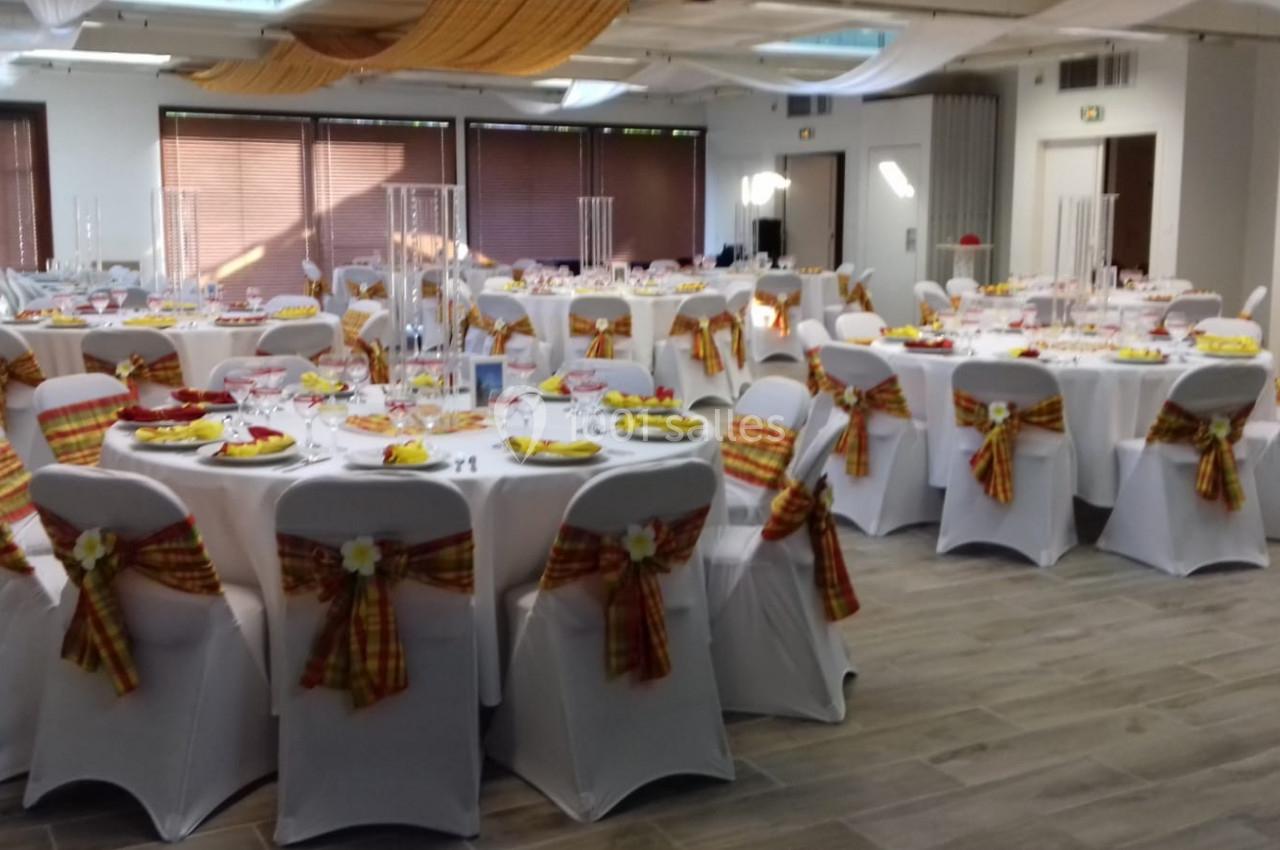 Salle de réception décorée avec des tables rondes, nappes blanches et chaises ornées de nœuds colorés.