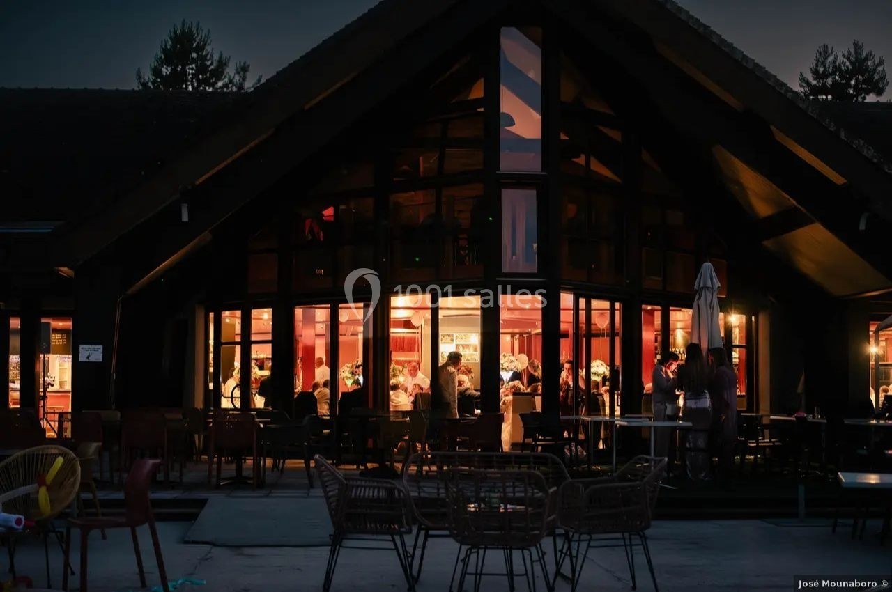 Vue nocturne d'un restaurant avec des baies vitrées illuminées, des clients visibles à l'intérieur et une terrasse sombre.