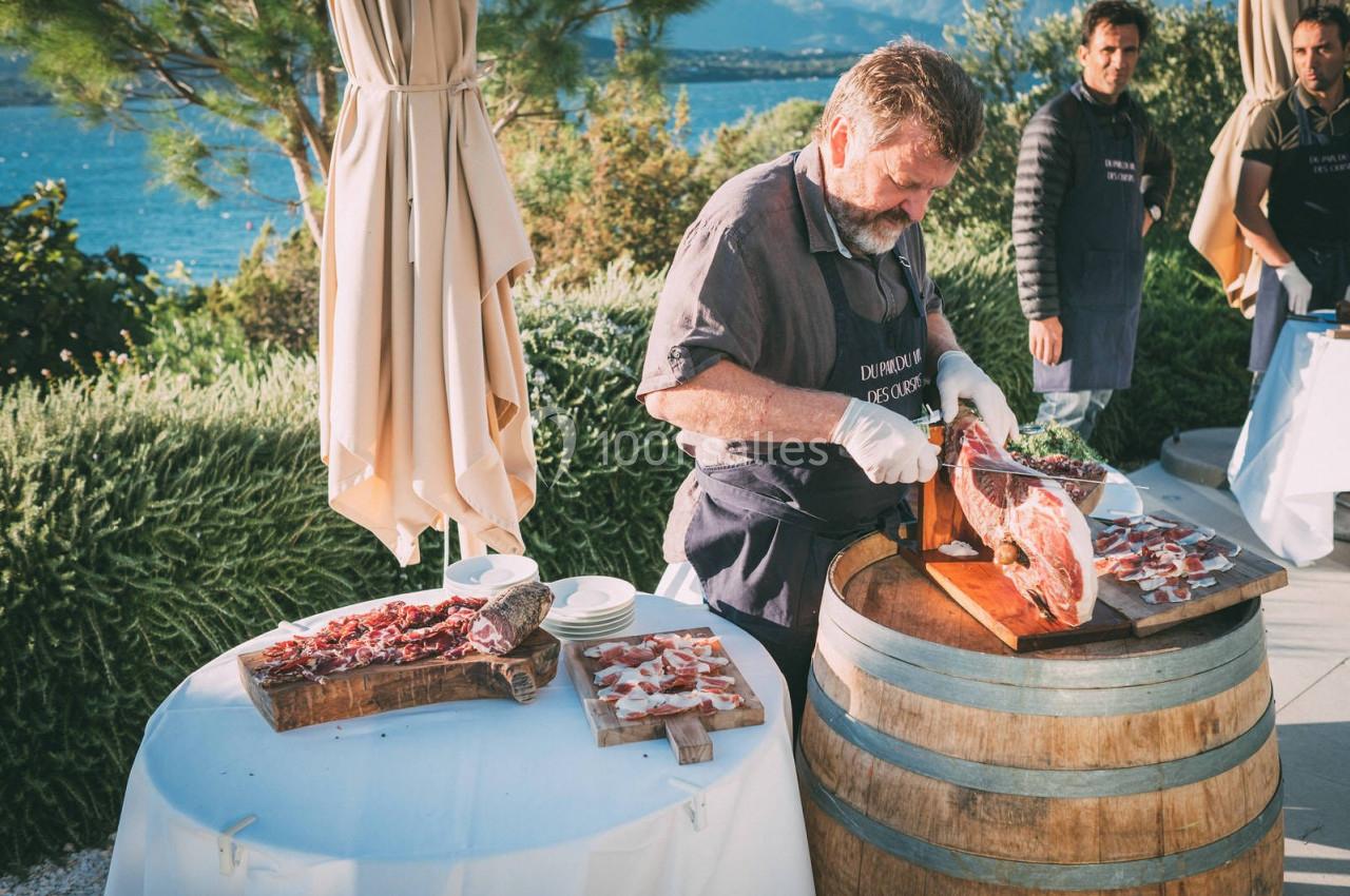 Un homme découpe du jambon sur un tonneau en bois lors d'un événement en extérieur avec vue sur la mer.