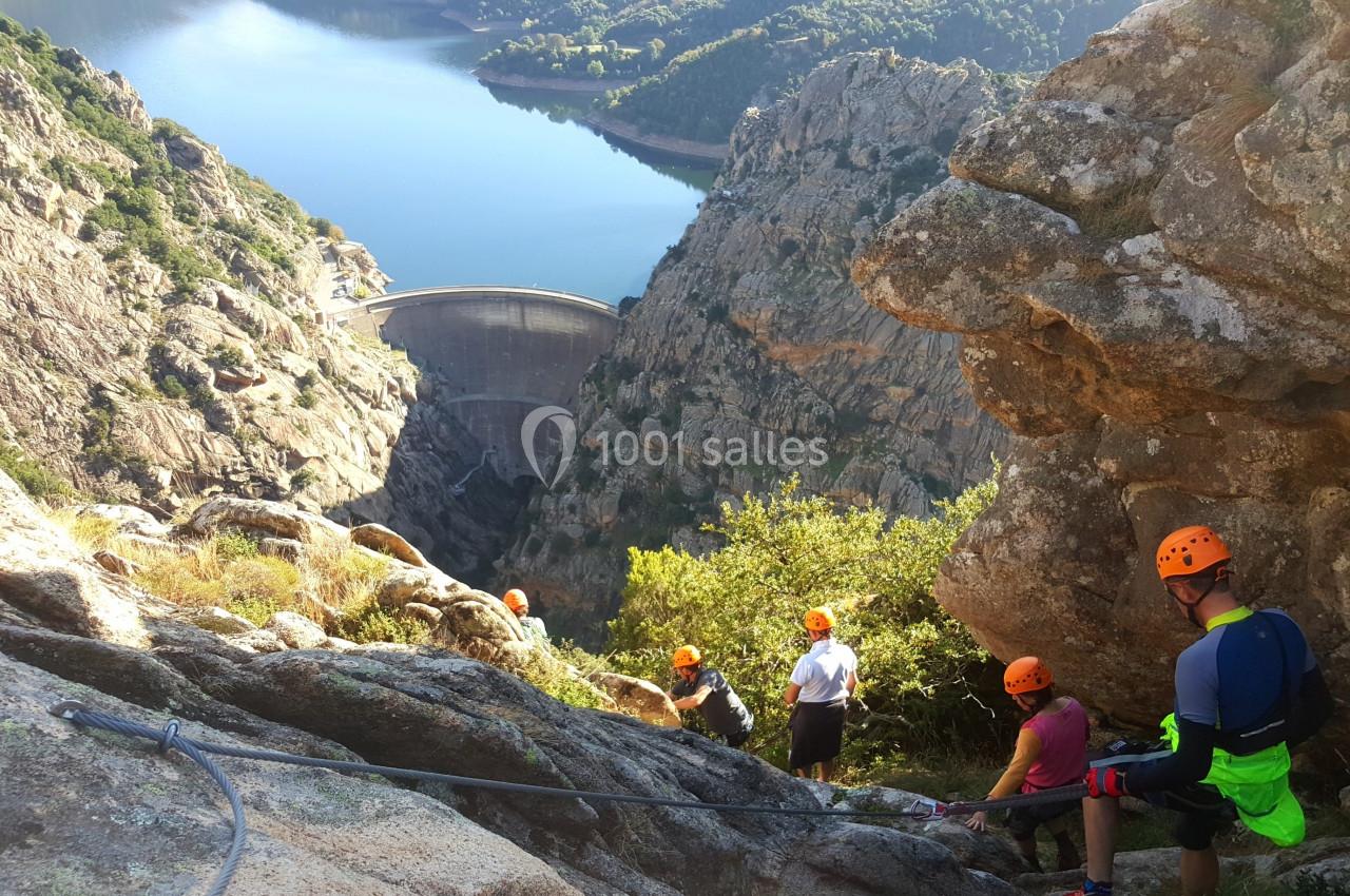 Des personnes équipées pour la descente en rappel dans un paysage montagneux avec un barrage visible au loin.