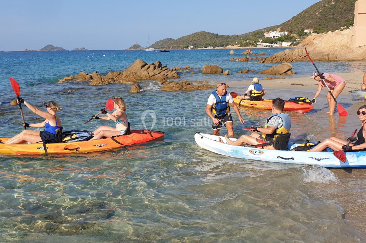Des personnes en kayak se préparent à naviguer depuis une plage bordée de rochers et d'eau claire, par temps ensoleillé.