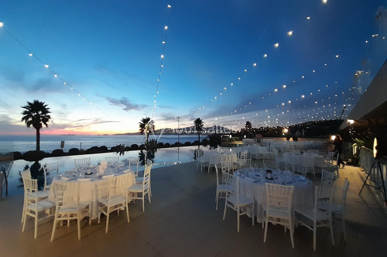 Tables dressées en extérieur près d'une piscine, sous des guirlandes lumineuses, au coucher du soleil.