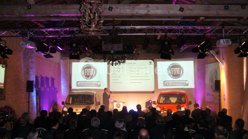 Un conférencier présente des véhicules Fiat devant un public dans une salle éclairée avec des projecteurs.