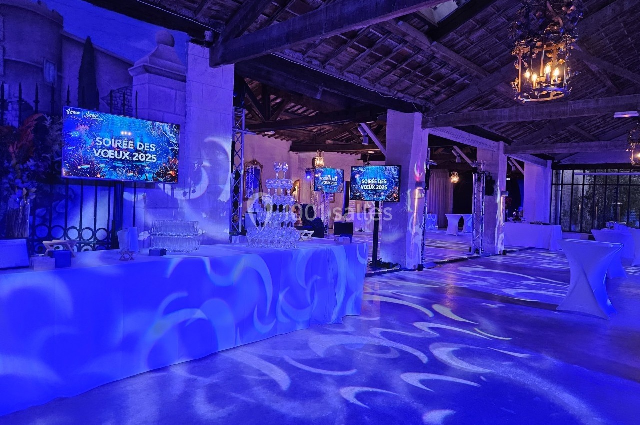 Salle décorée avec éclairage bleu, écrans affichant ’Soirée des Vœux 2025’, tables hautes et ambiance festive.