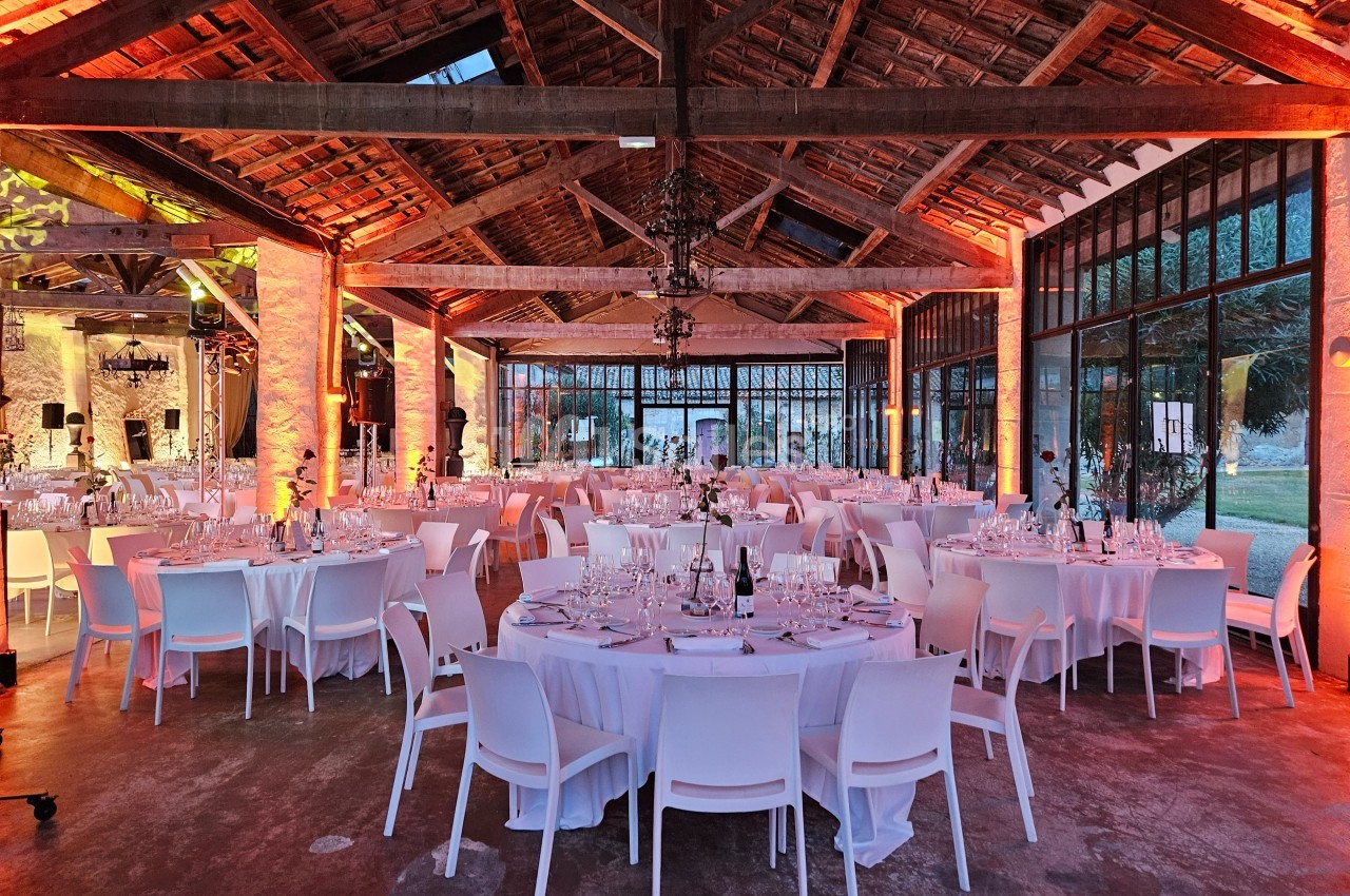 Salle de réception avec tables rondes dressées, chaises blanches et éclairage chaleureux sous une charpente en bois.