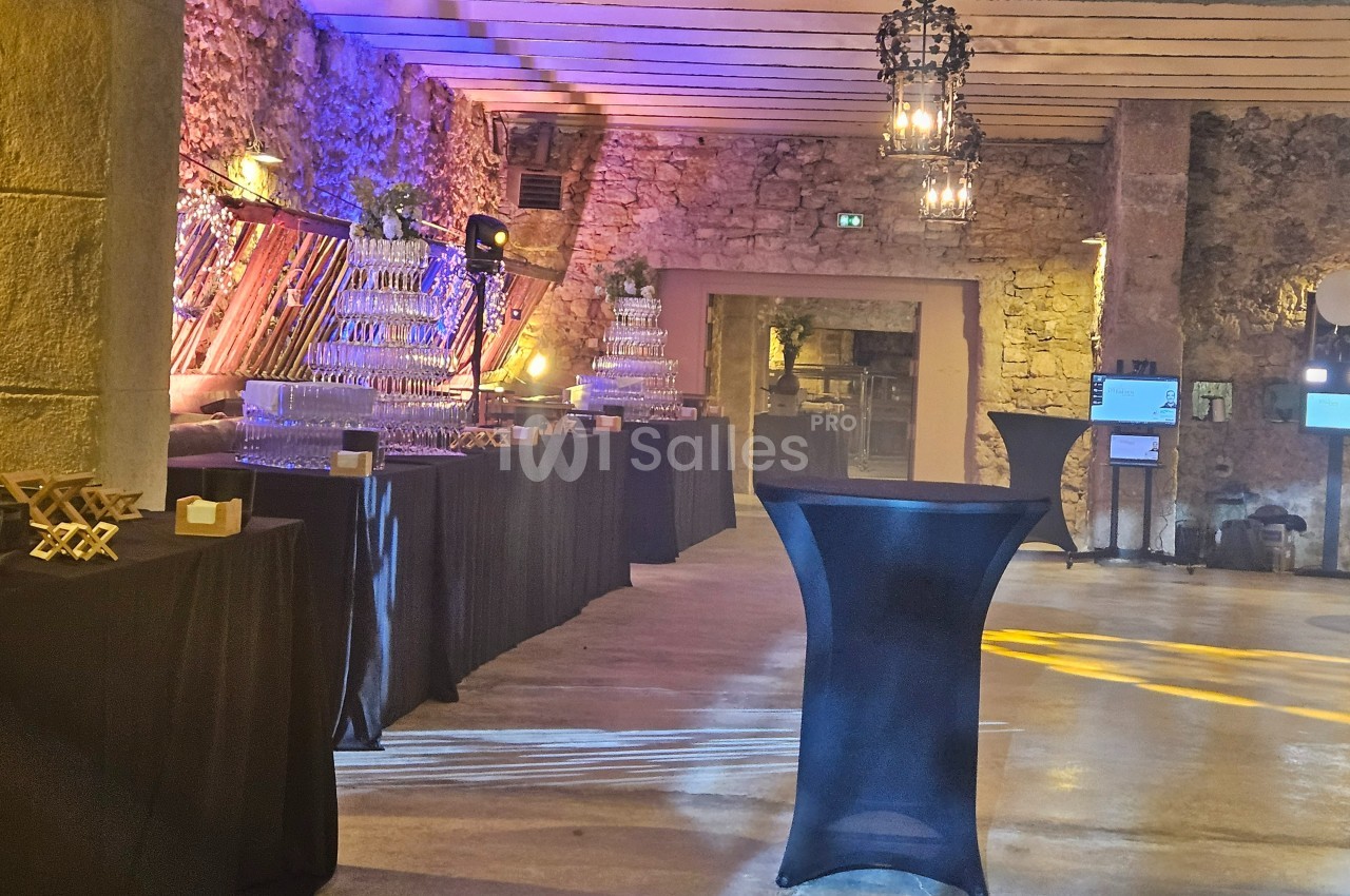 Salle en pierre avec éclairage chaleureux, tables hautes noires et buffet de verres empilés pour un événement.