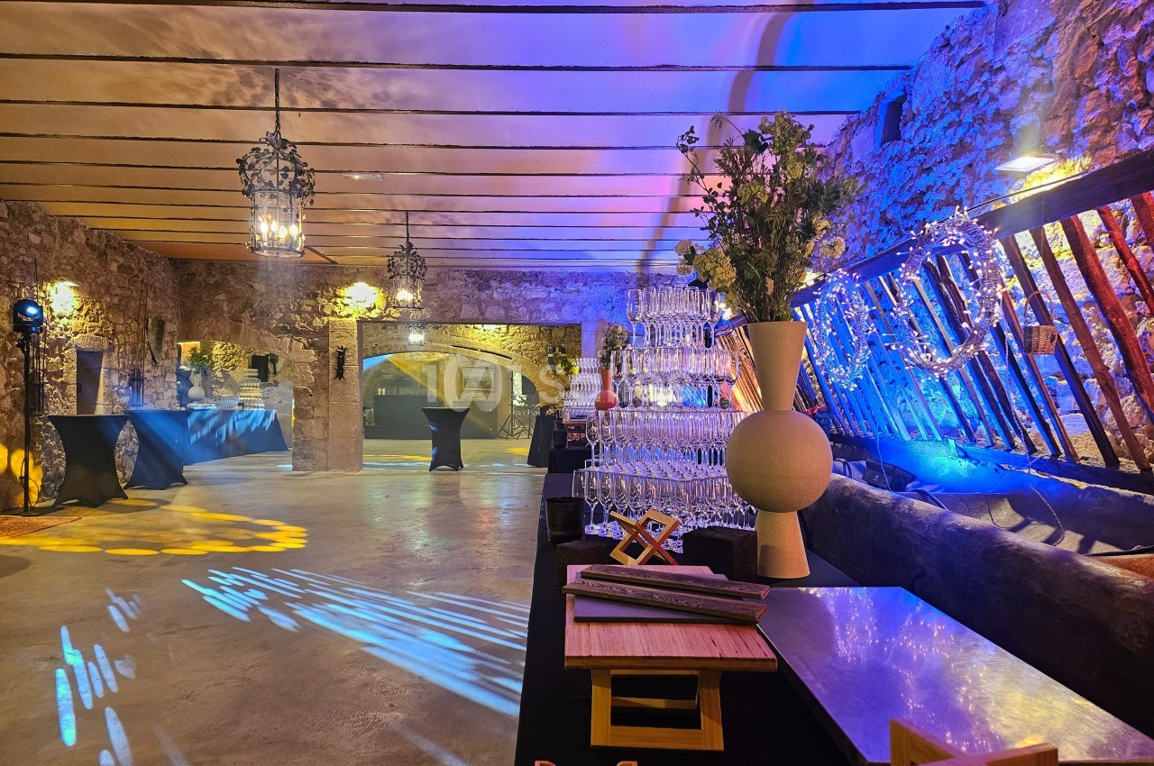 Salle en pierre éclairée avec des lumières colorées, tables hautes, décorations et pyramide de verres à champagne.