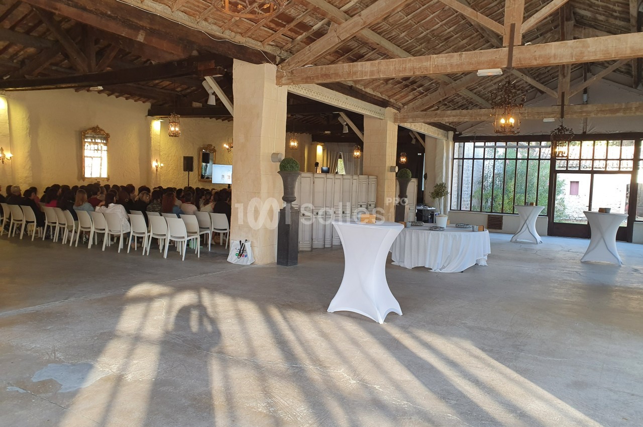 Salle spacieuse avec charpente en bois, tables hautes nappées et rangées de chaises occupées pour un événement.