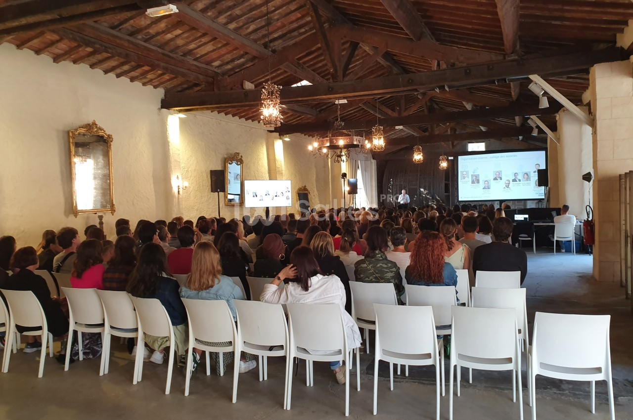 Un groupe de personnes assises dans une salle avec un plafond en bois, assistant à une présentation sur écran.