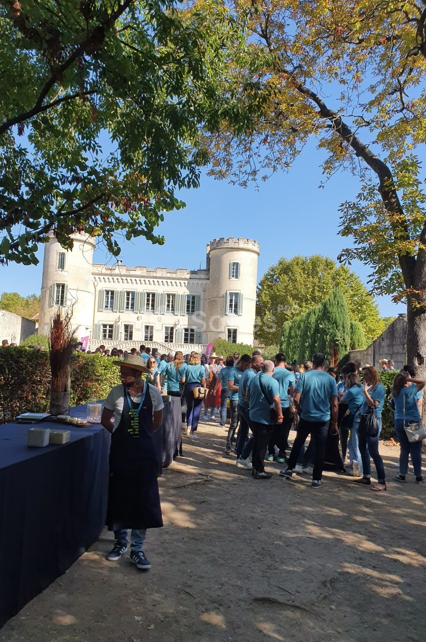 Groupe de personnes rassemblées devant un château entouré d'arbres, avec des tables dressées en extérieur.