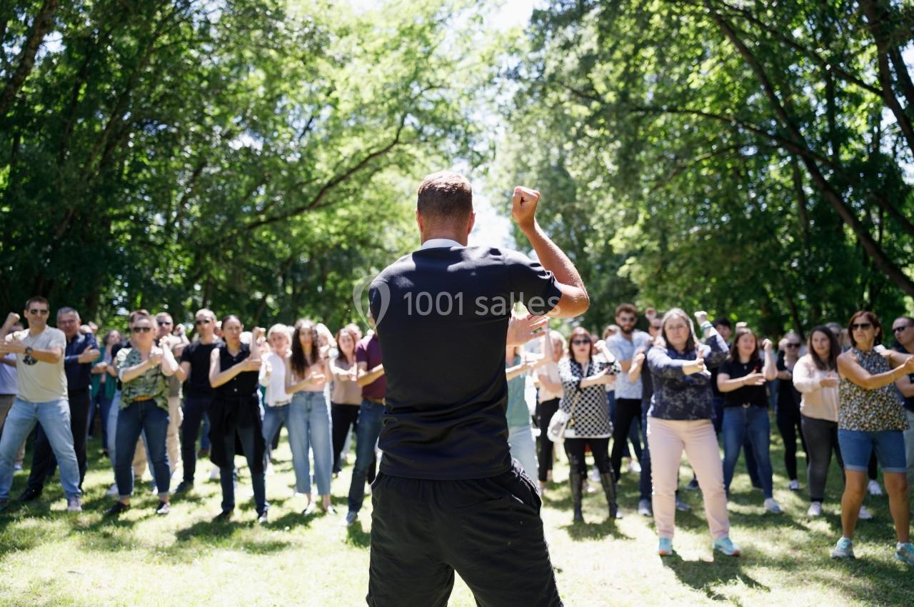 Un homme de dos anime un groupe de personnes rassemblées dans un parc en plein air, sous des arbres.