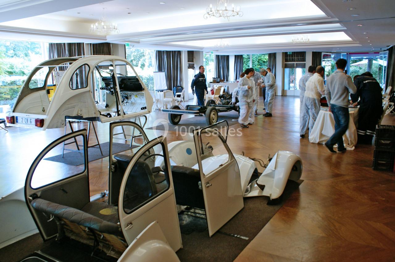 Des personnes assemblent des pièces de carrosserie de voitures dans une salle lumineuse avec parquet en bois.