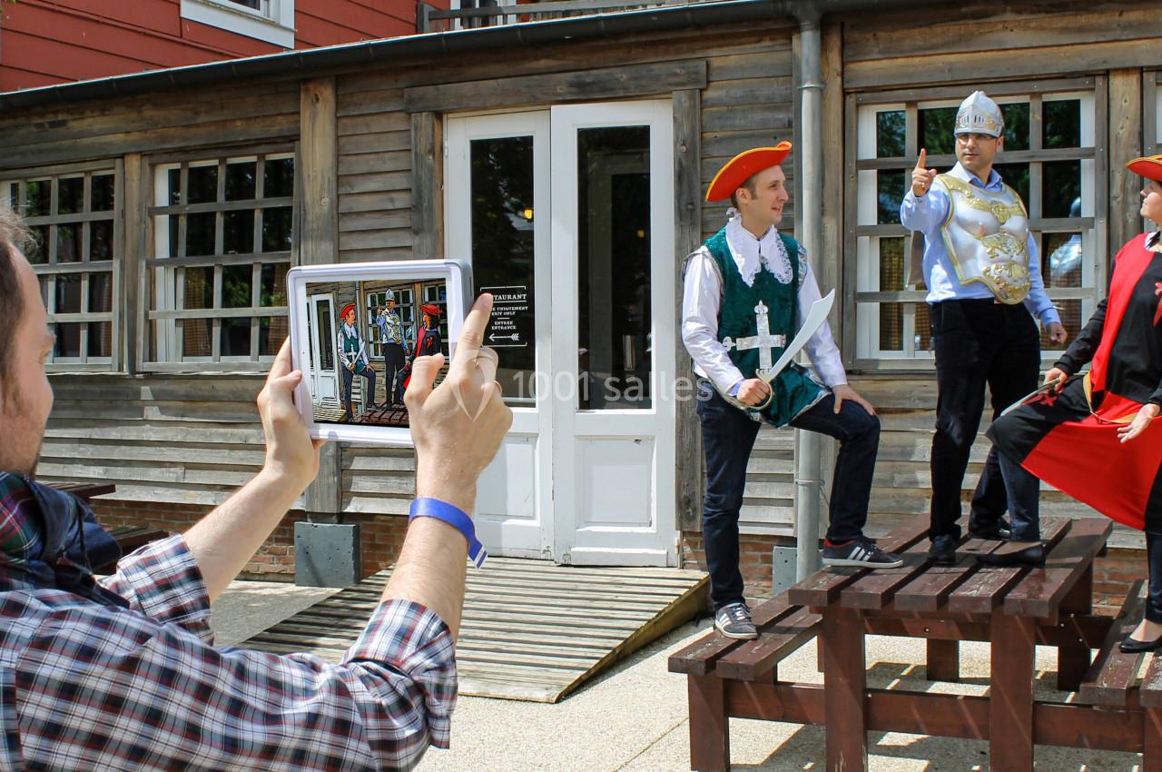 Un homme photographie trois personnes déguisées en costumes historiques posant sur des bancs devant un bâtiment en bois.
