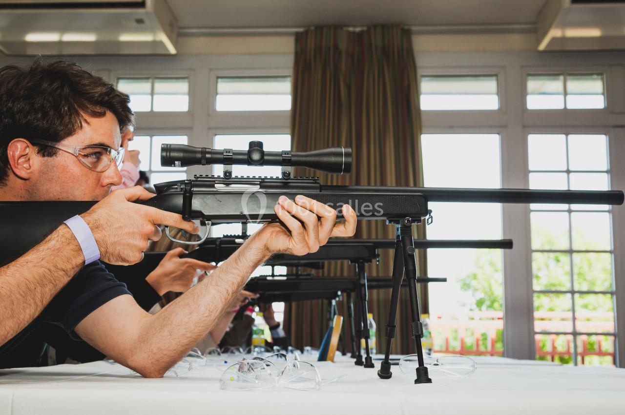Homme portant des lunettes de protection, visant avec une carabine équipée d'une lunette de visée dans une pièce lumineuse.