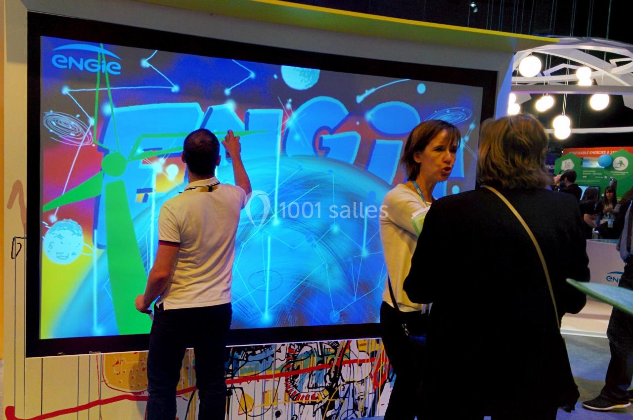 Trois personnes interagissent devant un écran tactile affichant des visuels colorés dans un espace d'exposition.