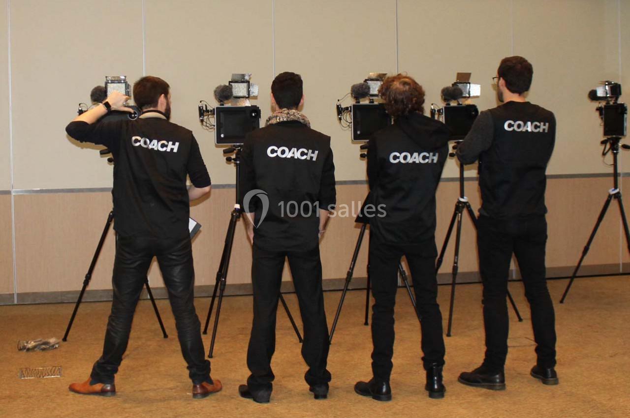 Quatre personnes de dos, portant des vêtements avec l'inscription ’COACH’, devant des caméras sur trépieds.