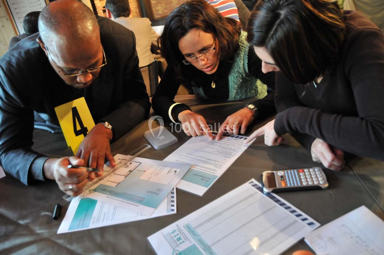 Trois personnes concentrées examinent des documents et des plans sur une table, avec une calculatrice à proximité.