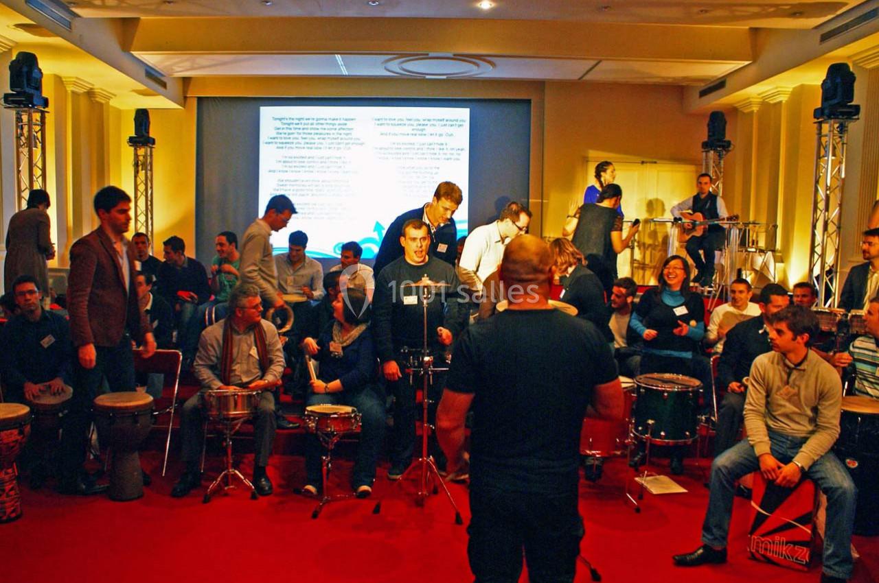 Un groupe de personnes joue des percussions dans une salle éclairée, devant un écran de projection.
