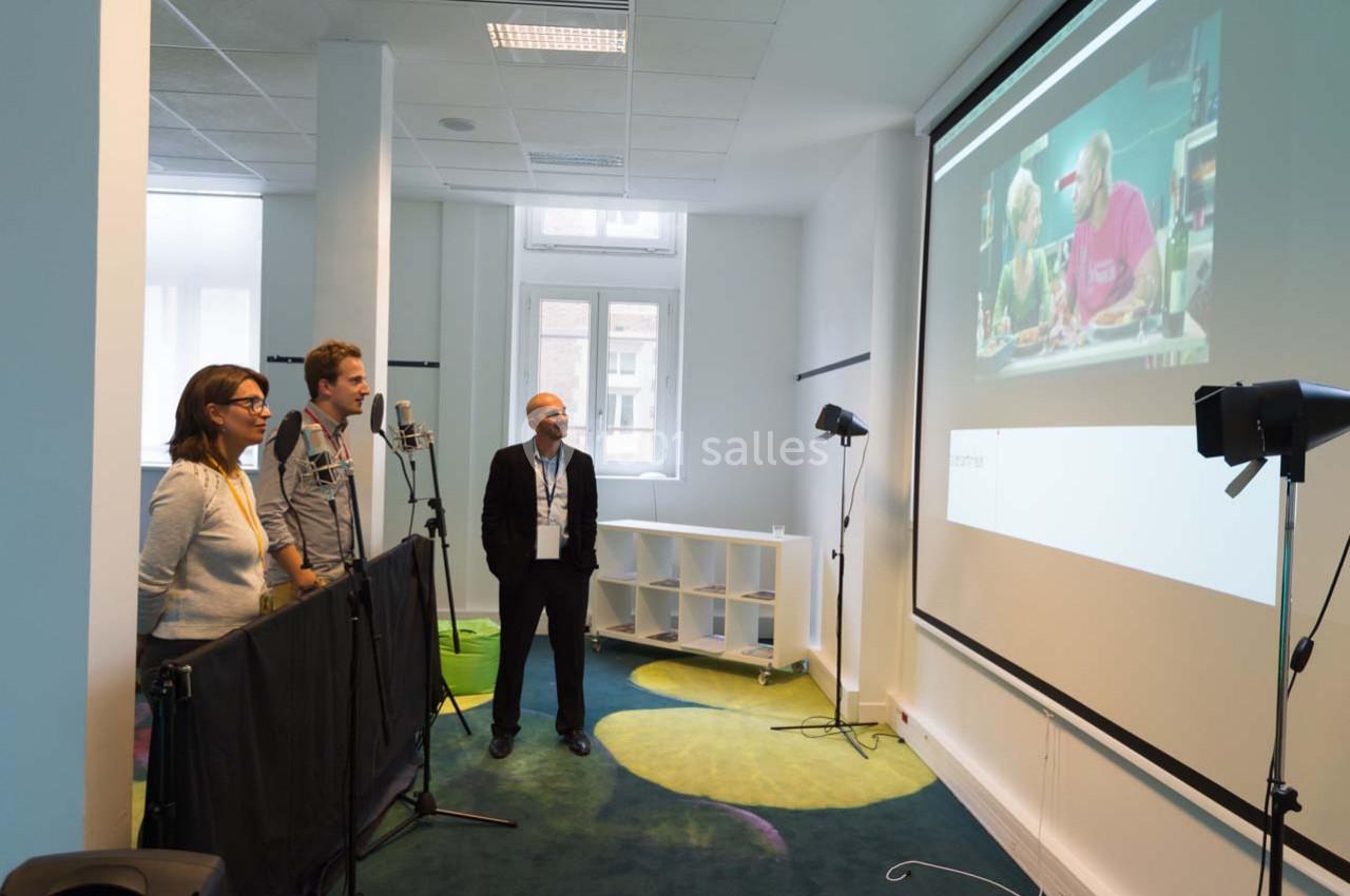 Trois personnes enregistrent des voix dans un studio équipé de micros, avec une projection vidéo en arrière-plan.
