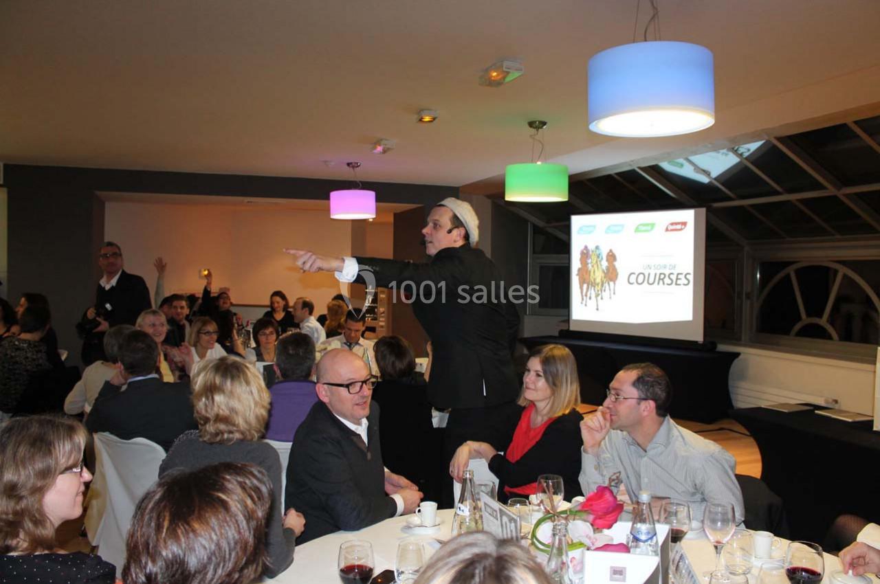 Un homme anime une soirée conviviale avec des participants assis autour de tables dans une salle éclairée.