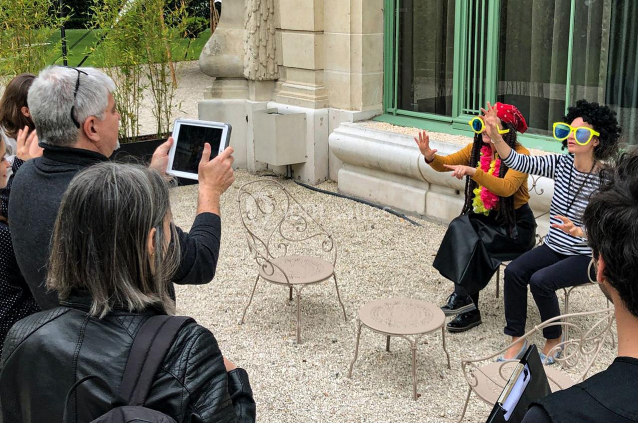 Des personnes photographient deux individus déguisés avec des lunettes colorées, assis sur des chaises en extérieur.