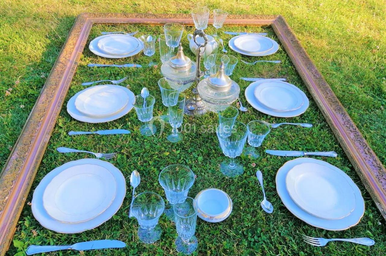 Table dressée sur de l'herbe avec vaisselle en porcelaine, couverts en métal et verres en cristal, encadrée par un cadre…