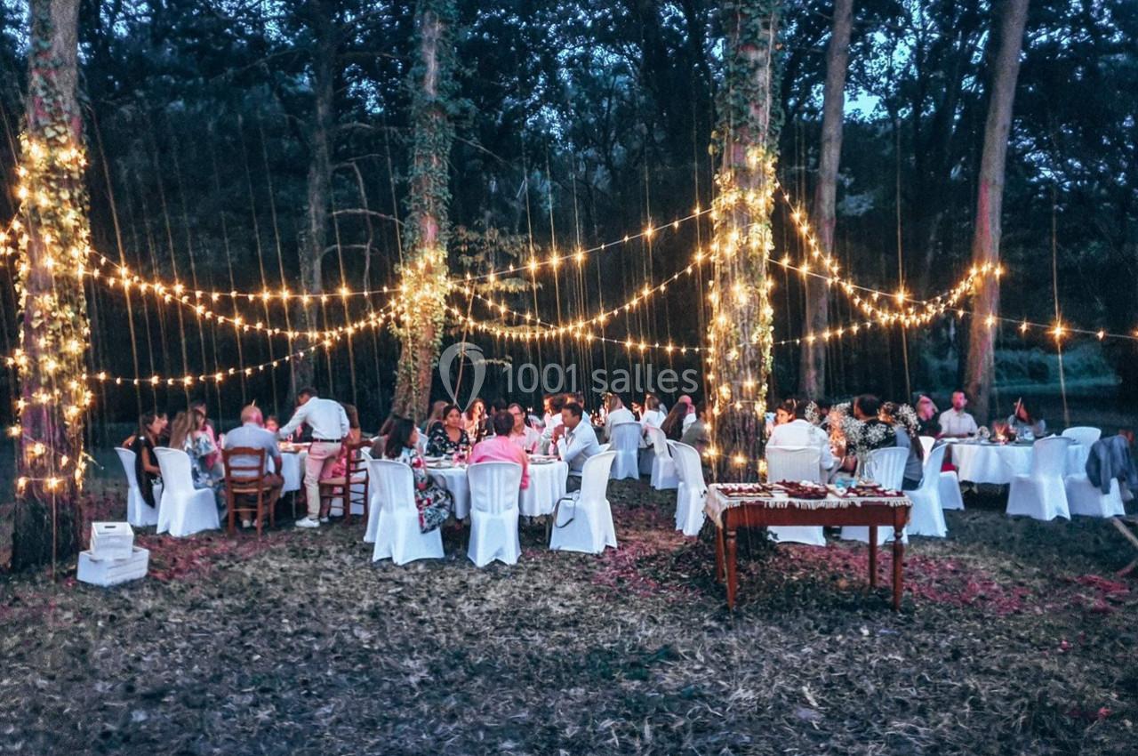 Des invités dînent en plein air sous des guirlandes lumineuses suspendues entre les arbres, à la tombée de la nuit.