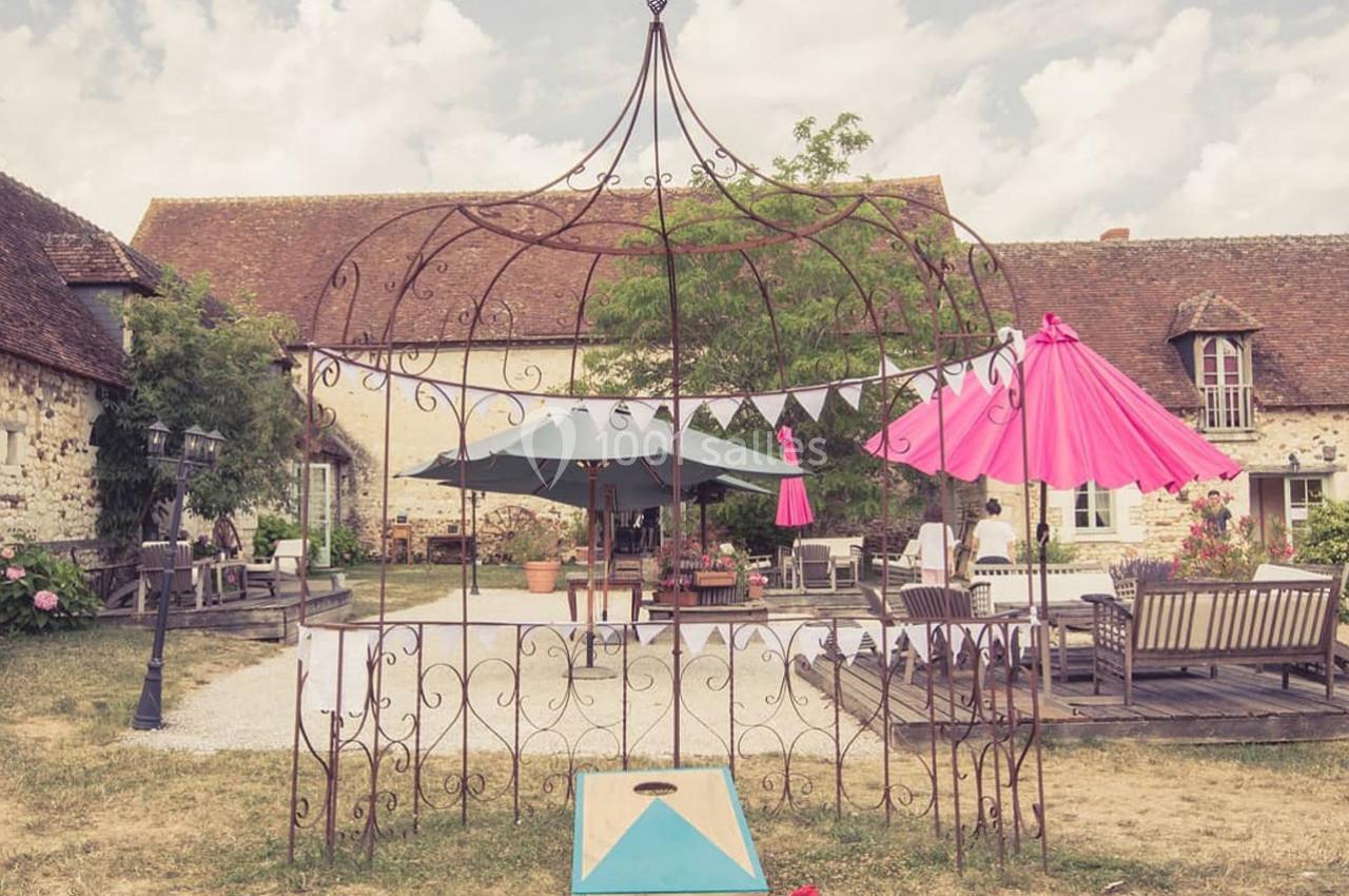 Cour extérieure avec mobilier de jardin, parasols colorés et structure métallique décorative devant une maison en pierre.