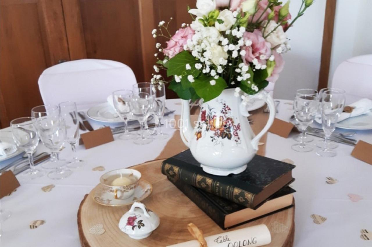 Centre de table avec bouquet de fleurs dans une théière, posé sur des livres et une rondelle de bois, entouré de vaisselle.