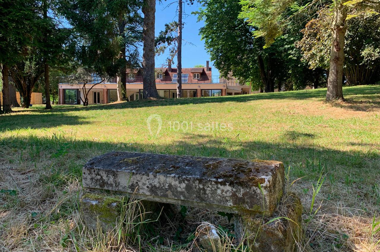 Banc en pierre ancienne dans un parc verdoyant, avec une maison moderne visible en arrière-plan.