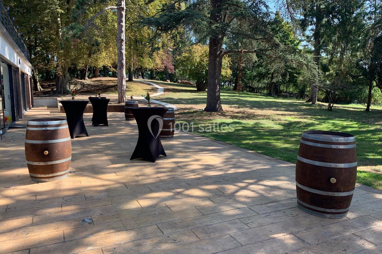 Terrasse en bois avec des tonneaux et des tables hautes noires, entourée d'arbres dans un cadre verdoyant.
