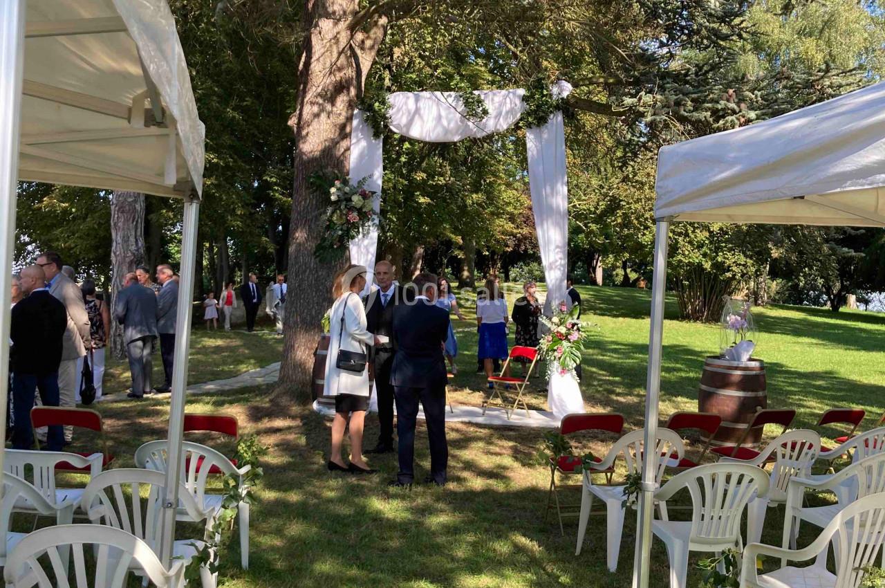 Cérémonie en plein air sous des arbres, avec des chaises blanches disposées et un arche décoré de tissu blanc.