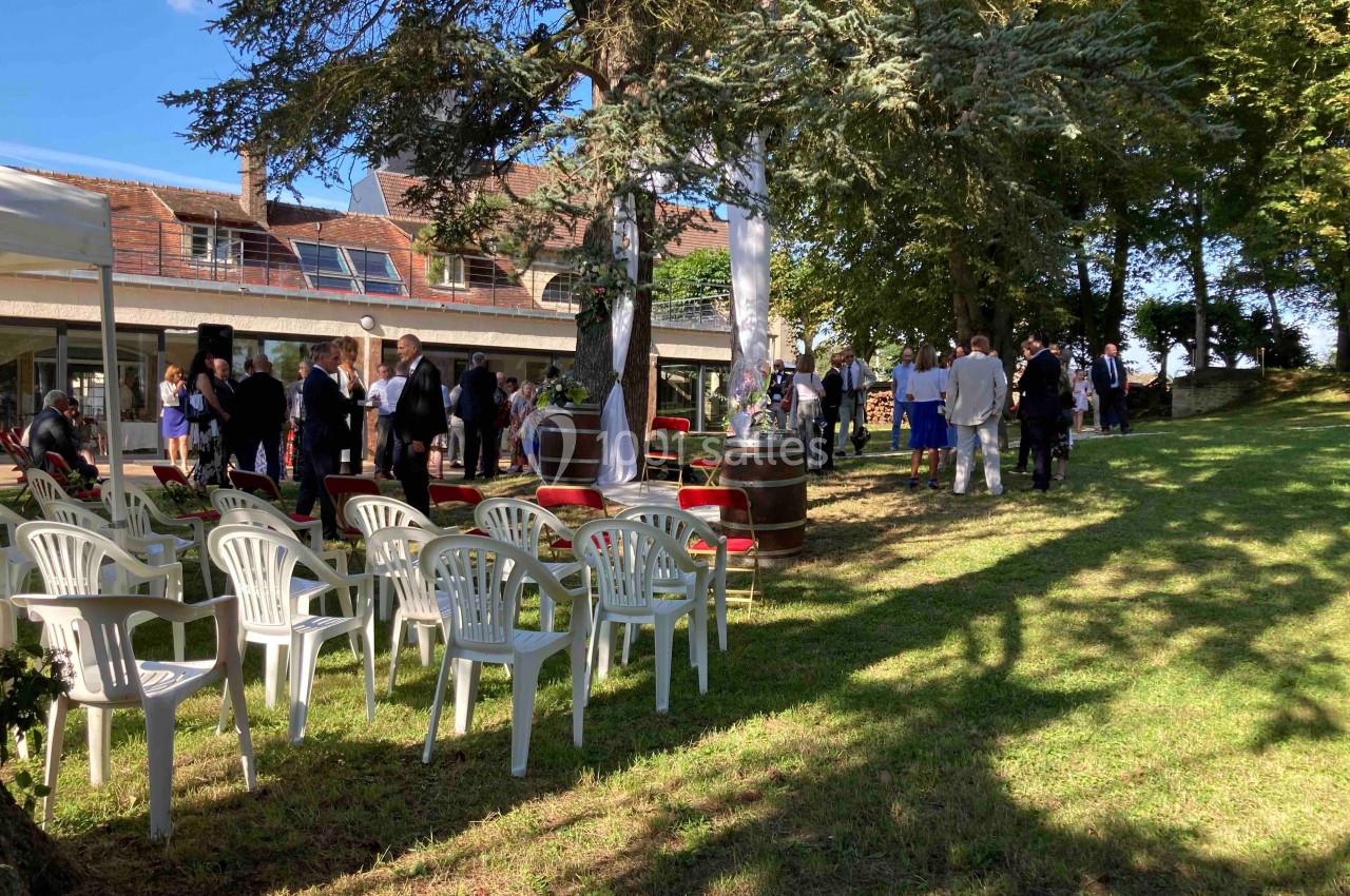 Des personnes rassemblées dans un jardin ombragé avec des chaises en plastique blanc et un bâtiment en arrière-plan.