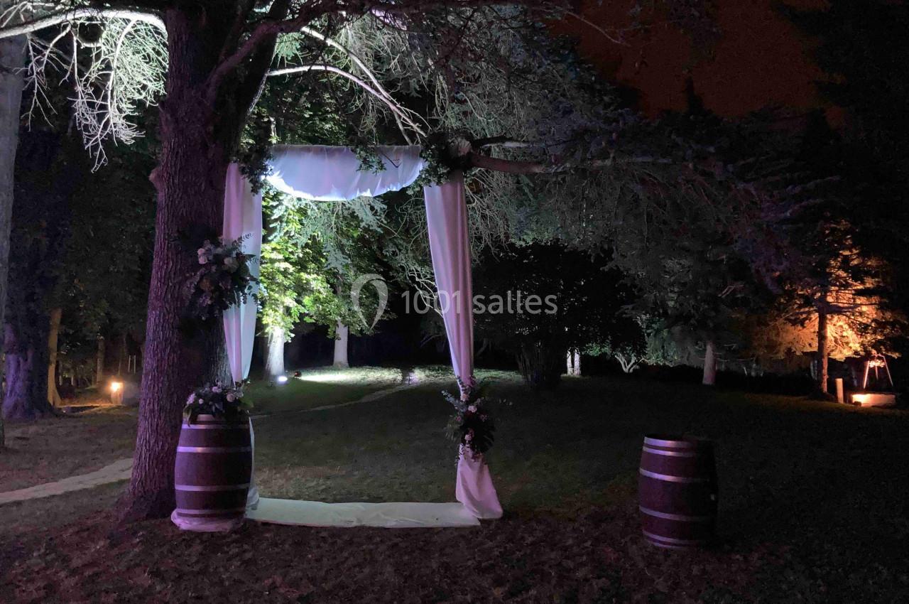 Arche de mariage décorée de voilages blancs et de fleurs, installée en extérieur sous des arbres éclairés la nuit.