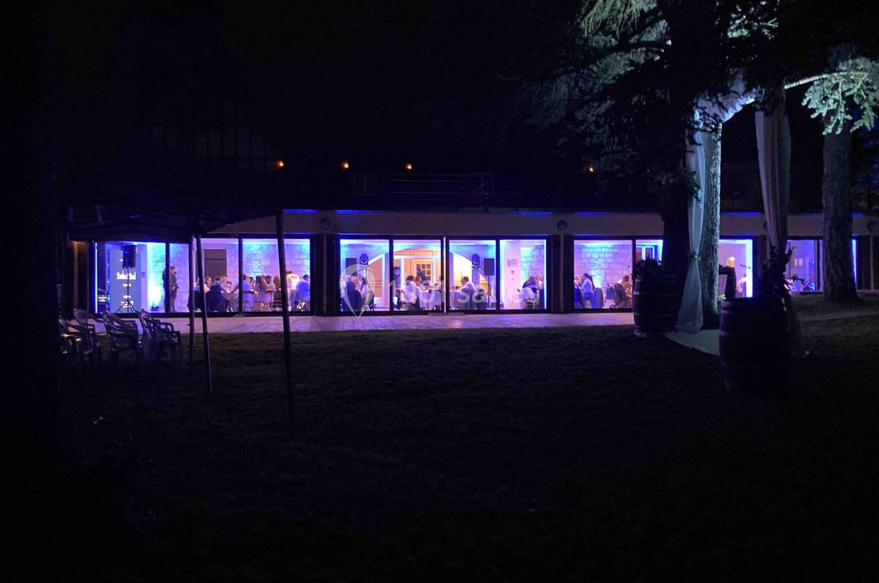 Salle vitrée éclairée en bleu la nuit, avec des personnes visibles à l'intérieur et des arbres en premier plan.