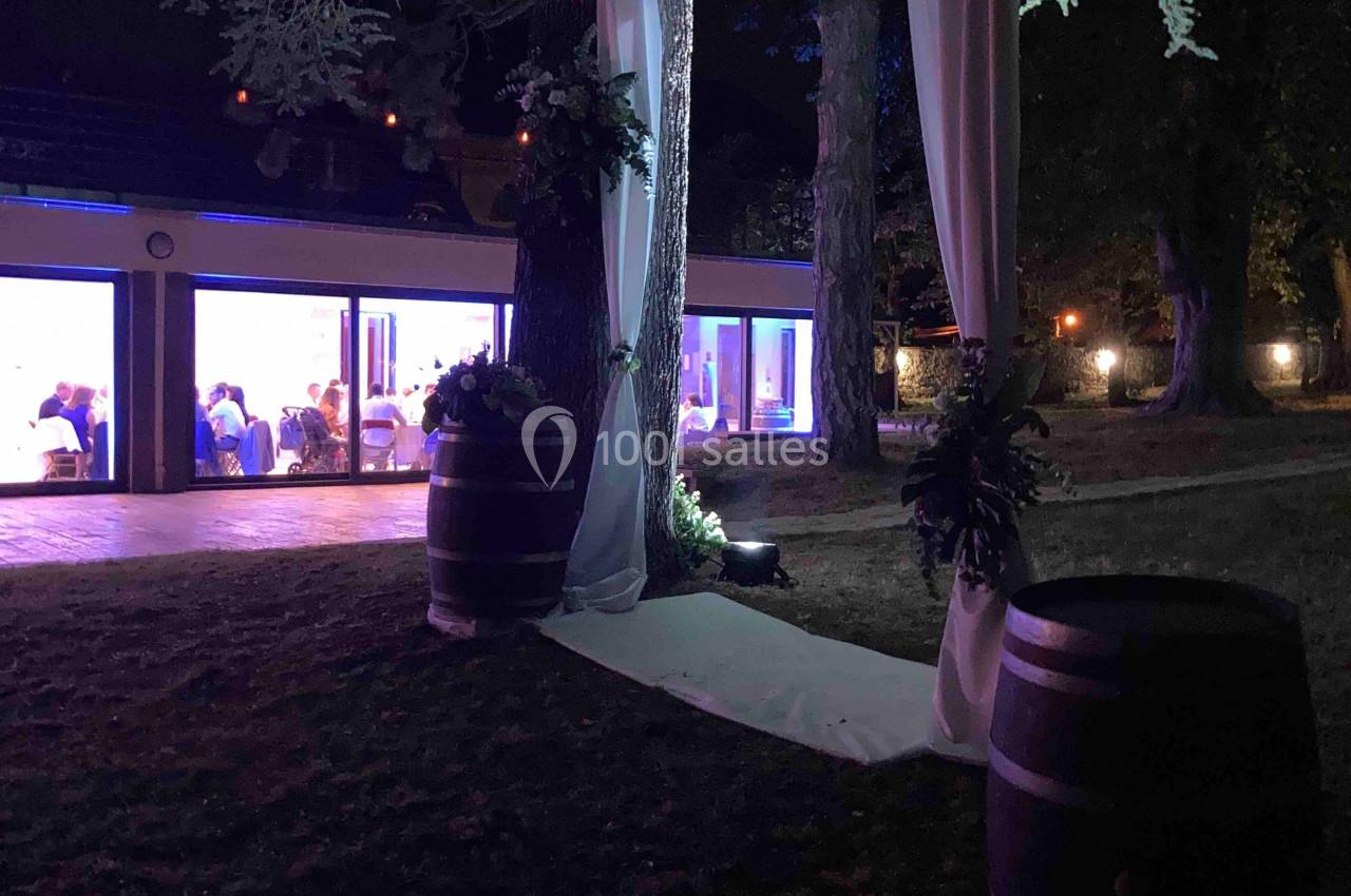 Décoration de mariage en extérieur avec arche lumineuse sous un arbre, barils en bois et salle éclairée en arrière-plan.