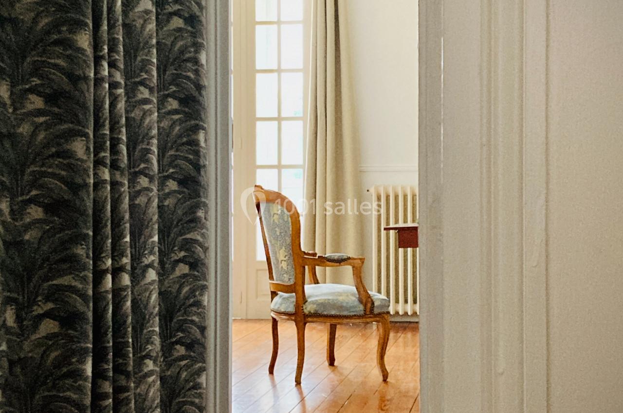 Une chaise en bois avec assise rembourrée est placée près d'une fenêtre dans une pièce lumineuse au sol en parquet.