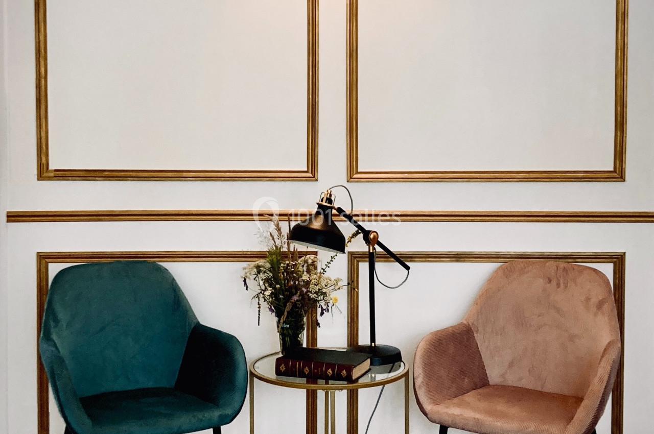 Deux fauteuils colorés entourent une table ronde avec une lampe, des livres et un bouquet, sur un sol en damier.