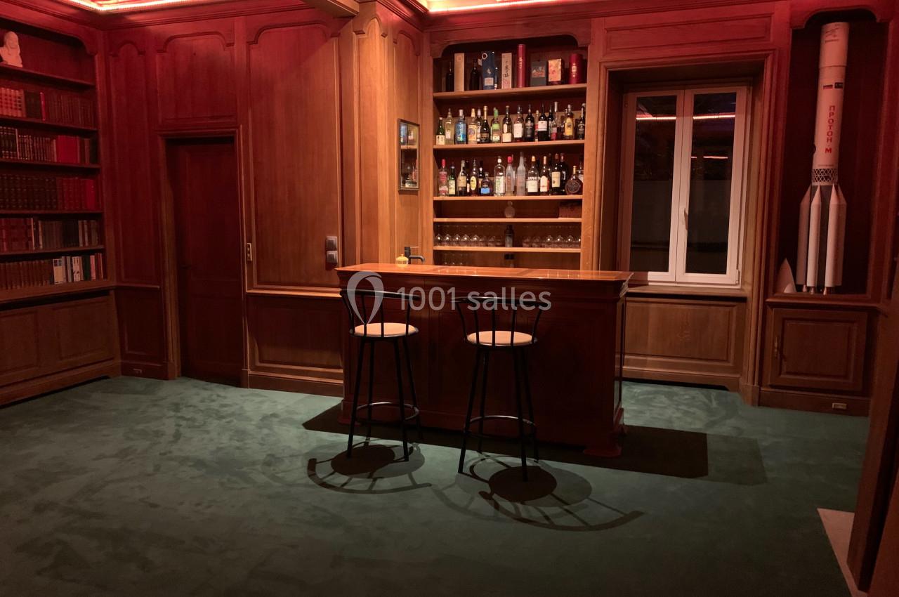 Bar en bois avec étagères remplies de bouteilles, deux tabourets et une décoration de fusée dans un intérieur chaleureux.