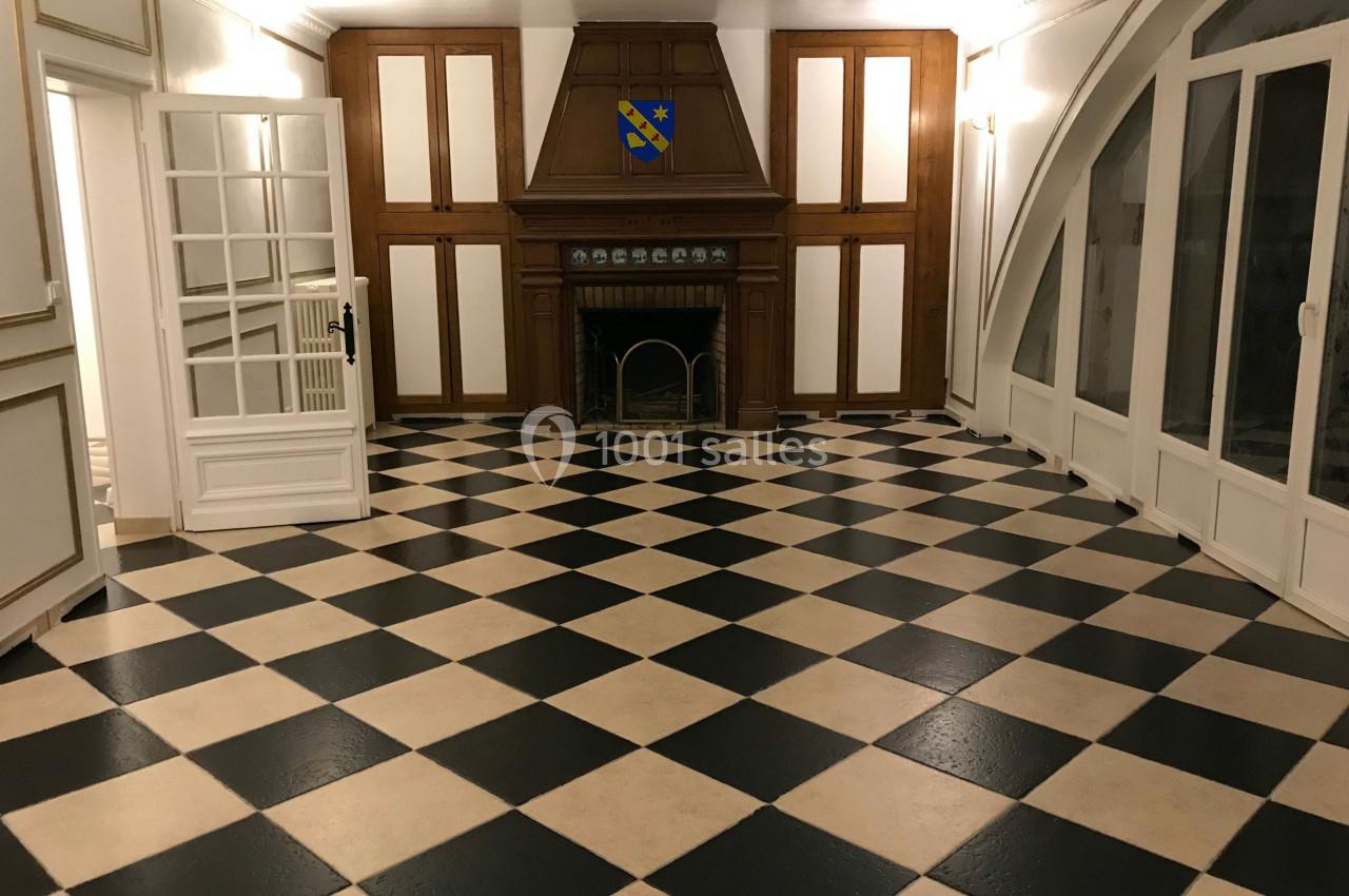 Salle avec un sol en damier noir et beige, cheminée en bois, murs blancs et grandes fenêtres cintrées.