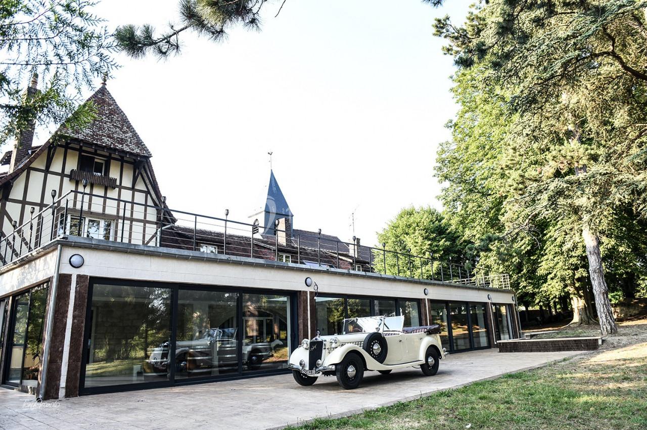 Voiture ancienne blanche stationnée devant un bâtiment à colombages entouré de verdure.