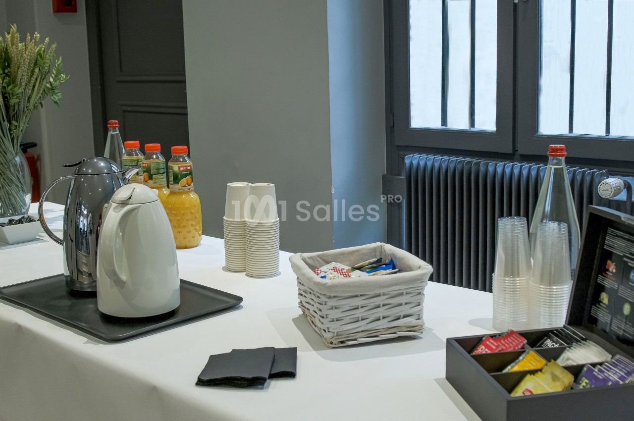 Table avec boissons, gobelets, thé, café, jus d'orange et accessoires, disposée près d'une fenêtre.