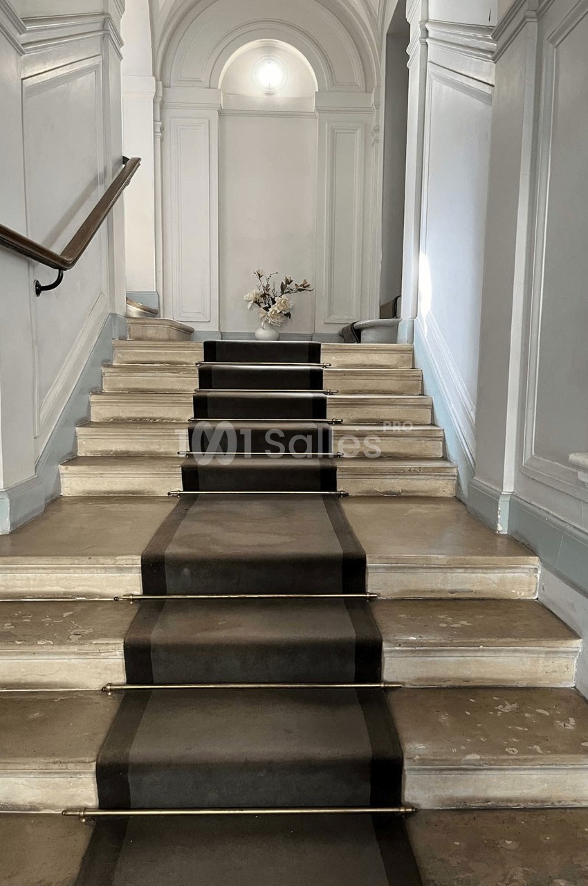 Escalier en pierre avec tapis central sombre, rampe en bois à gauche et vase de fleurs en arrière-plan.