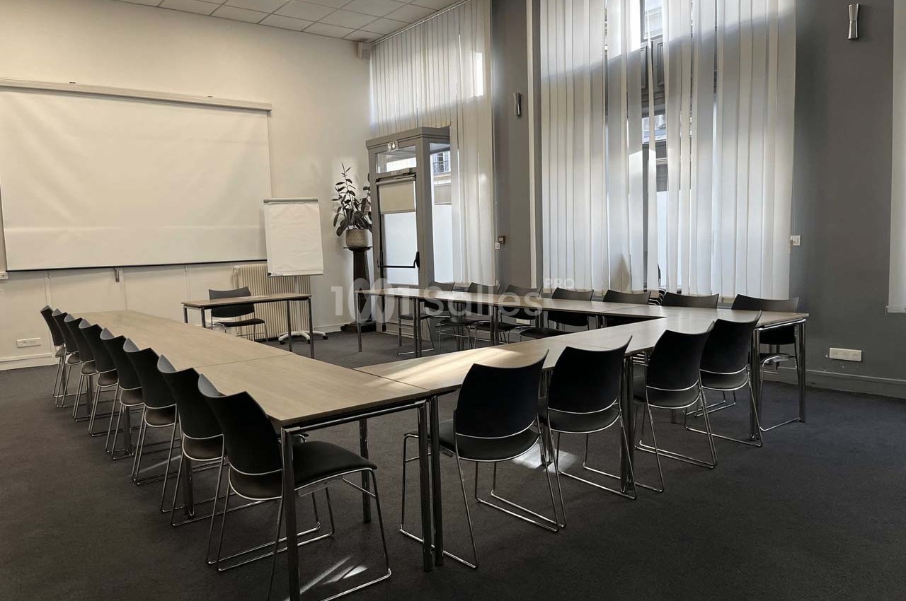 Salle de réunion lumineuse avec tables disposées en U, chaises noires et écran de projection.