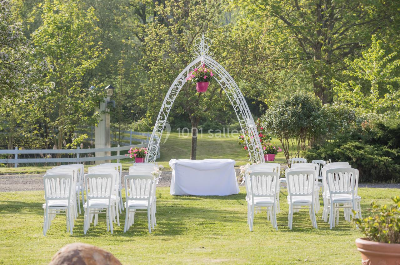 Chaises blanches disposées en rangées devant une arche décorée de fleurs dans un jardin verdoyant.